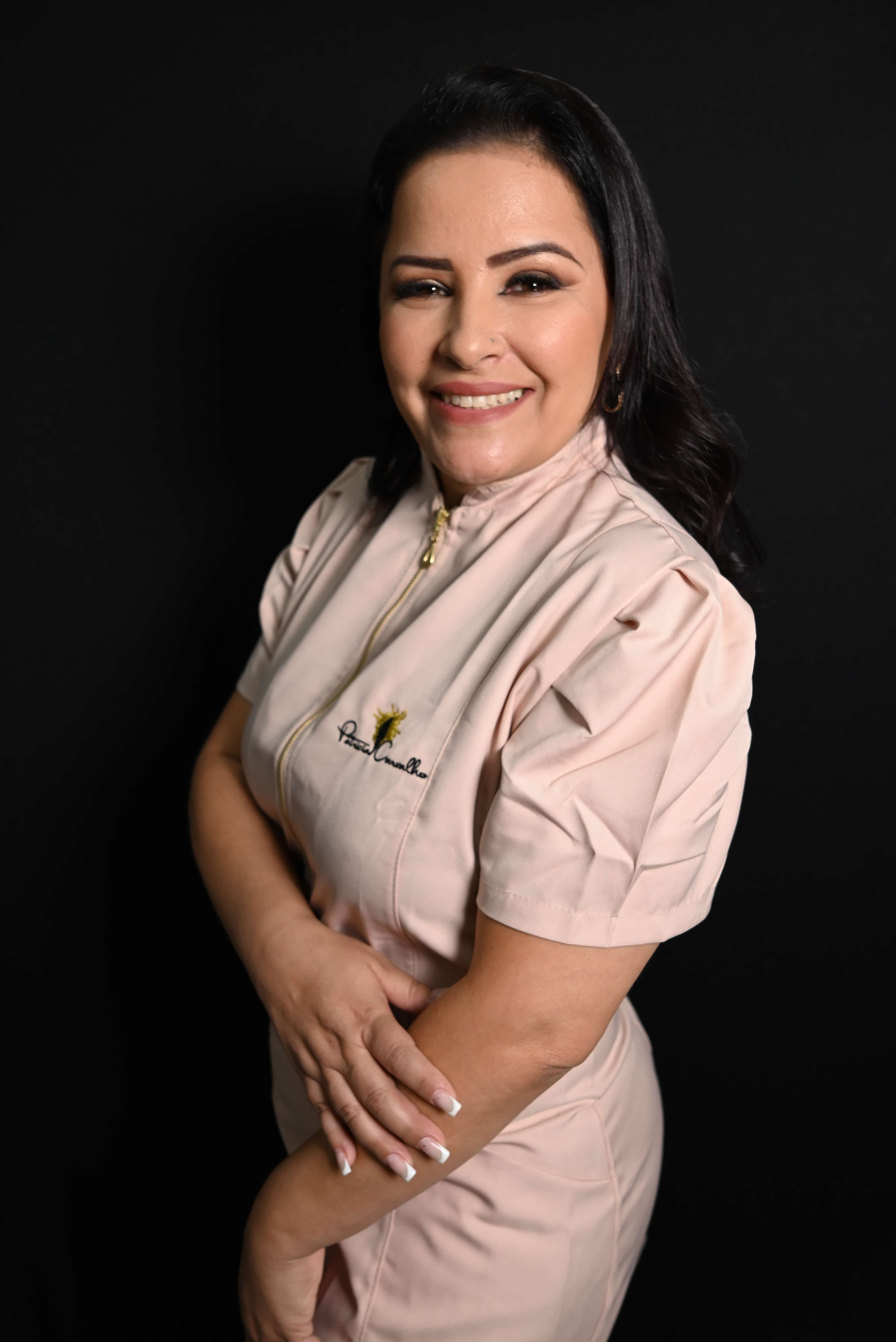 Retrato profissional de Patrícia Carvalho com uniforme bege, sorrindo de braços cruzados